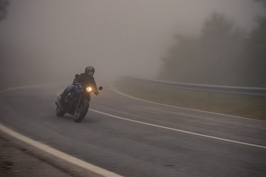 Un motorista circula entre la niebla por una carretera de curvas, con el faro encendido al amanecer